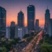 Ein fotorealistisches Bild zeigt ein geschäftiges Finanzviertel in Jakarta, Indonesien, bei Sonnenuntergang. Die moderne Skyline, dominiert von Wolkenkratzern, spiegelt die lebhaften Farben der untergehenden Sonne wider. Im Vordergrund steht das indonesische Börsengebäude, das für wirtschaftliches Wachstum steht. Belebte Straßen mit Menschen und Fahrzeugen vermitteln dynamische wirtschaftliche Aktivität. Zwischen den Gebäuden sind üppiges Grün und tropische Pflanzen zu sehen, die die natürliche Schönheit Indonesiens hervorheben. Die Szene fängt den Optimismus und die positive Dynamik der indonesischen Wirtschaft ein, beeinflusst durch kürzlich erfolgte Regierungsreformen und die Kürzung der Parlamentszuschüsse.
